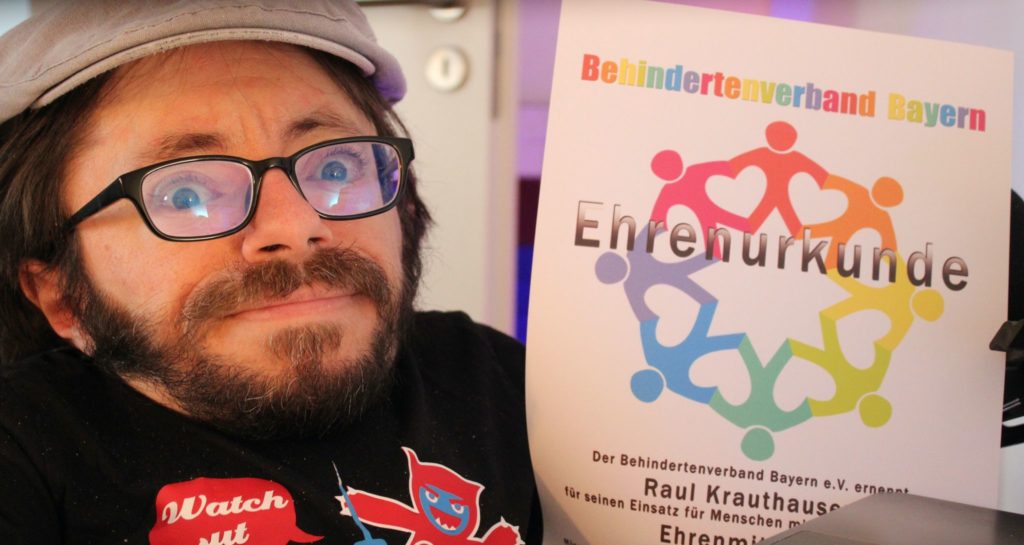 Auf dem Foto sieht man Raul Krauthausen mit seiner Ehrenurkunde vom Behindertenverband Bayern e.V.