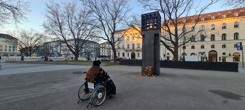 Rollstuhlfahrerin vor dem Mahnmal auf dem PLatz der Opfer des Nationalsozialismus. An das Mahnmal ist ein Krank gelehnt.